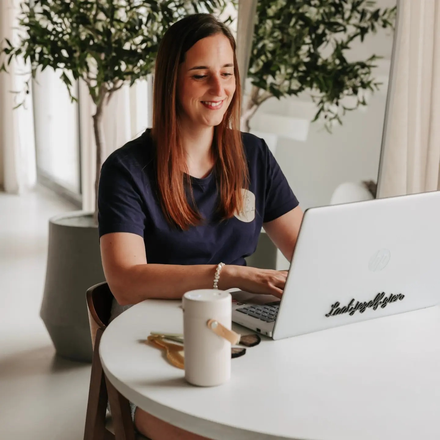 Daisy aan tafel achter haar laptop in een lichte ruimte met olijfboompjes